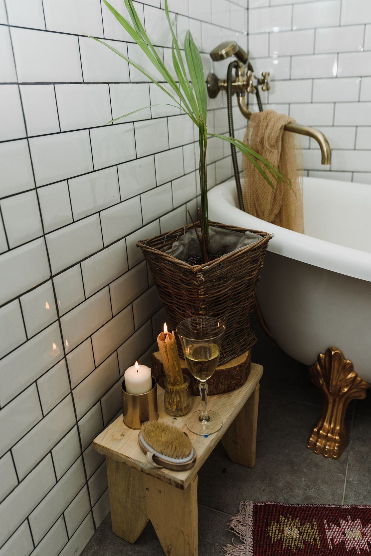 Wooden stool with lit candles in brass holders, a woven basket planter and a brass clawfoot tub in a cozy southwestern bathroom corner