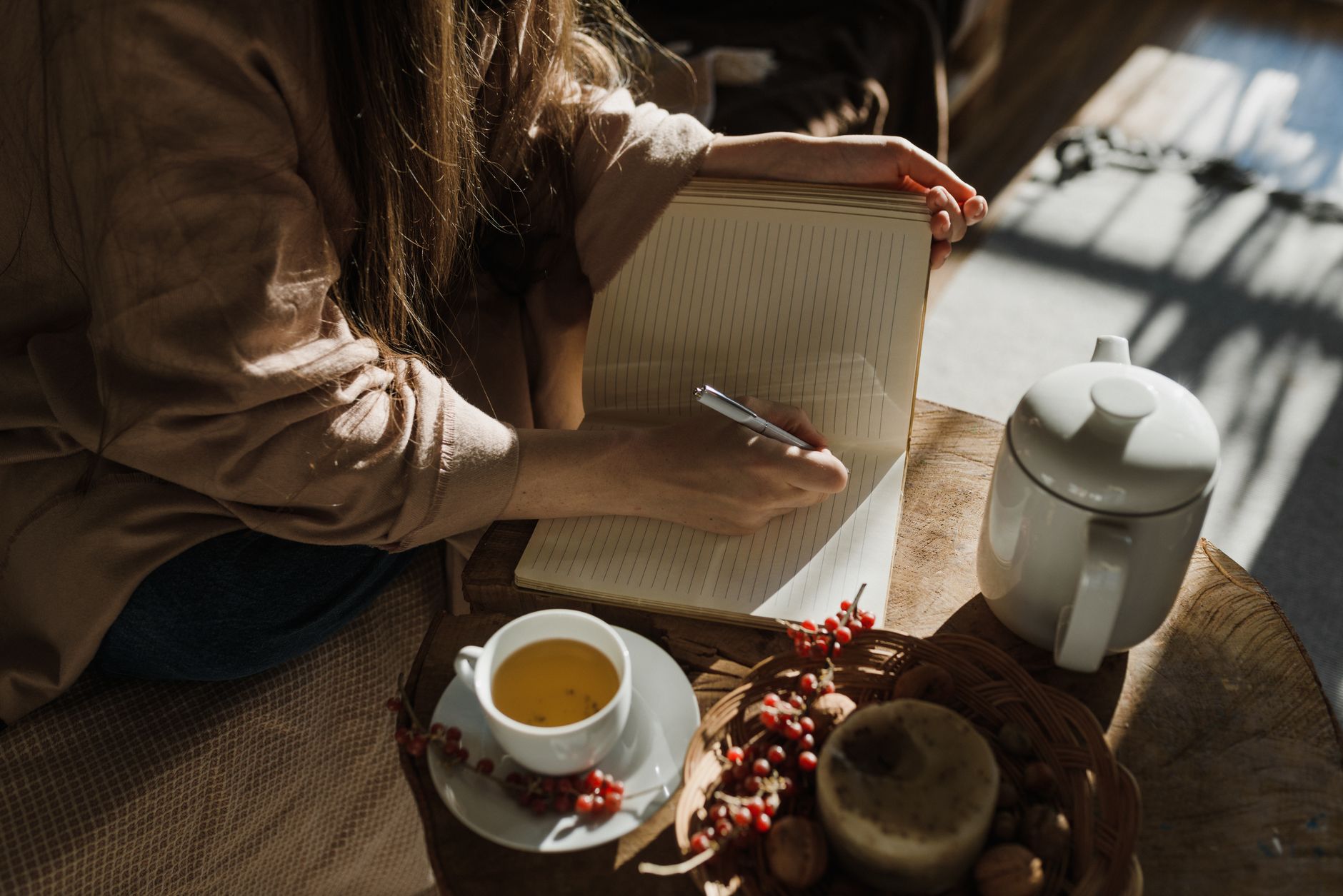 Seseorang menulis di buku harian di samping secangkir teh, dalam cahaya pagi yang lembut