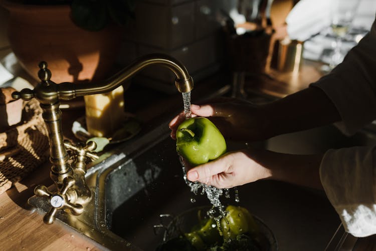 A Person Washing A Green Apple In A Sink With Running Water