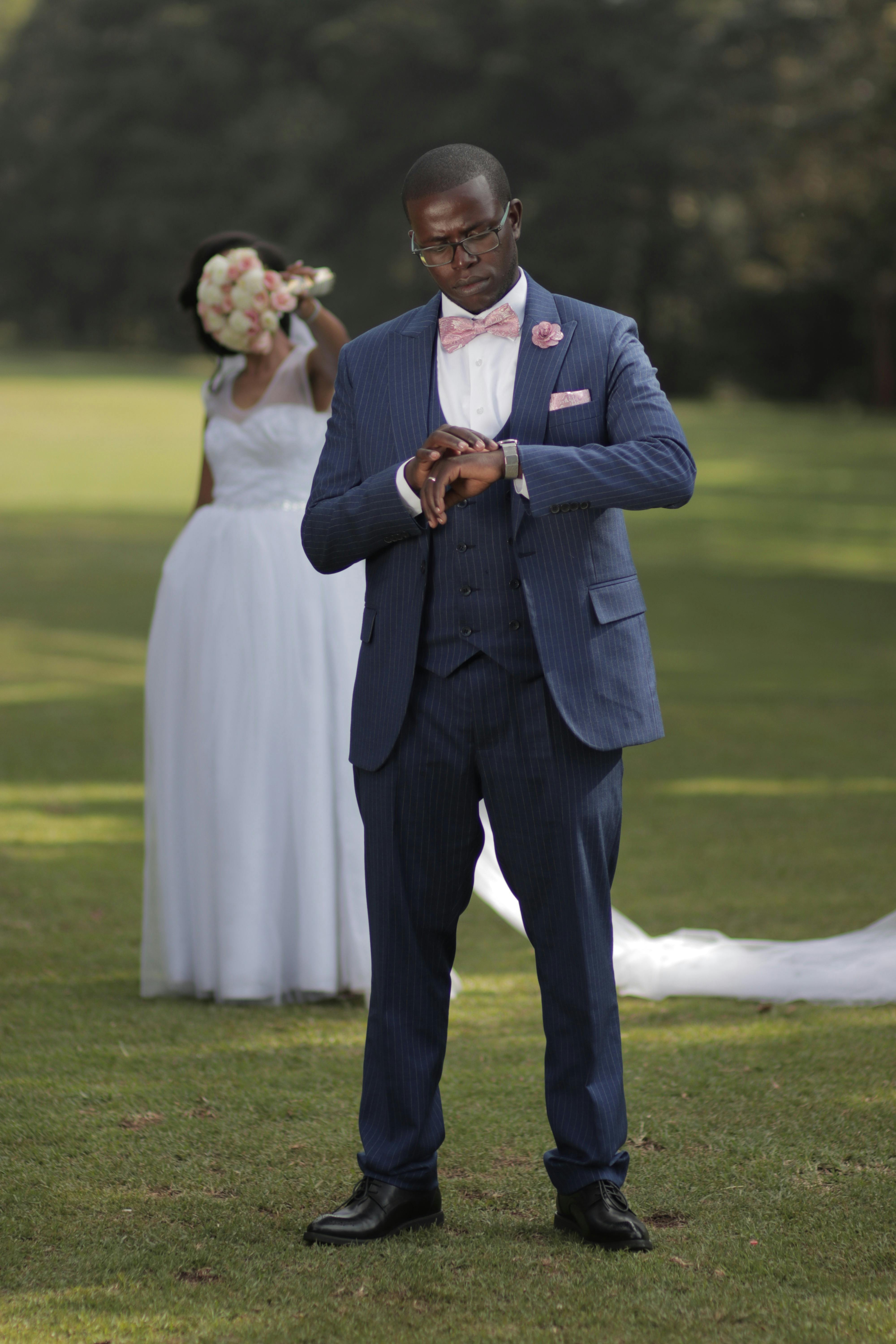 Groom in blue suit checking time outdoors with bride in the background.