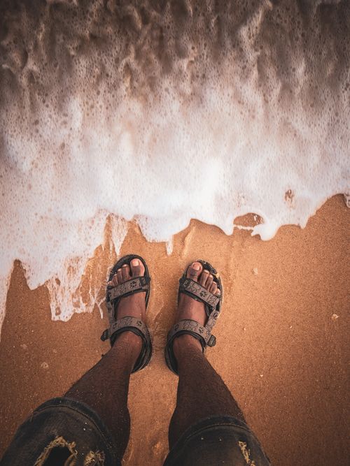 Free A person's feet wearing sandals on a sandy beach with sea foam gently touching the shore. Stock Photo