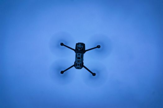 A drone seen flying in a clear blue sky, captured from below with motion blur on the rotors.