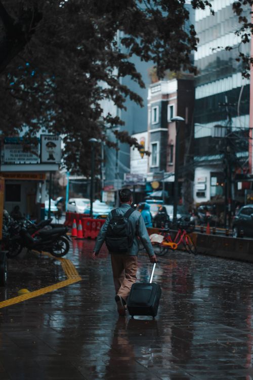 Free A man with a backpack pulls luggage on a wet city street in Jakarta, Indonesia. Stock Photo