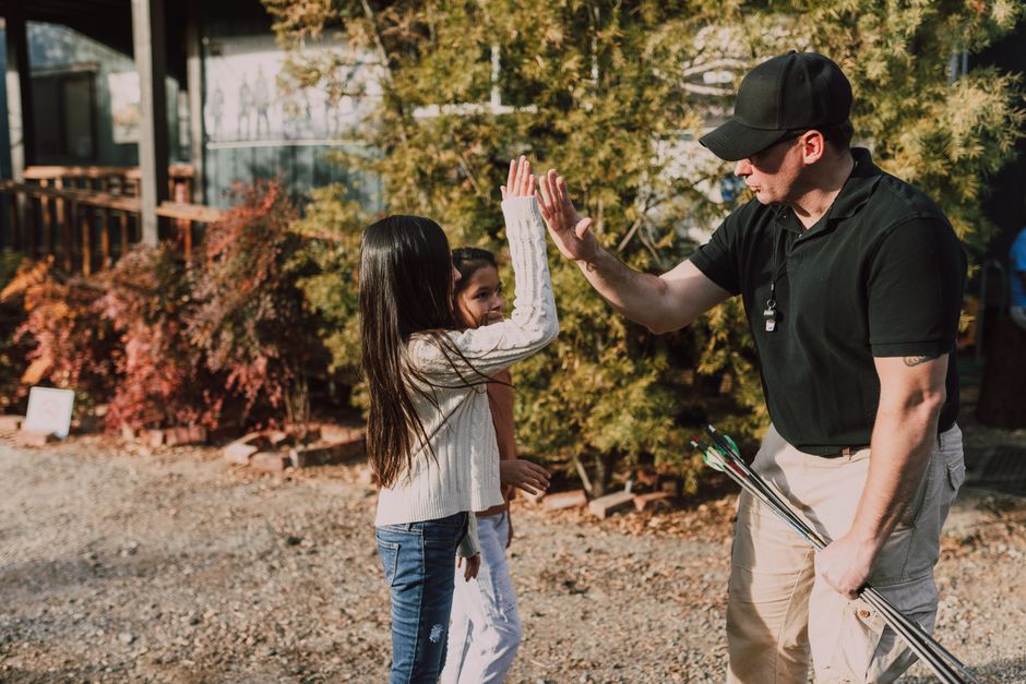 An instructor high-fiving a child at an archery range during a sunny day.