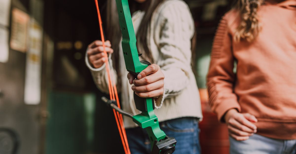 Two young girls engaged in archery practice with a vibrant green bow outdoors.