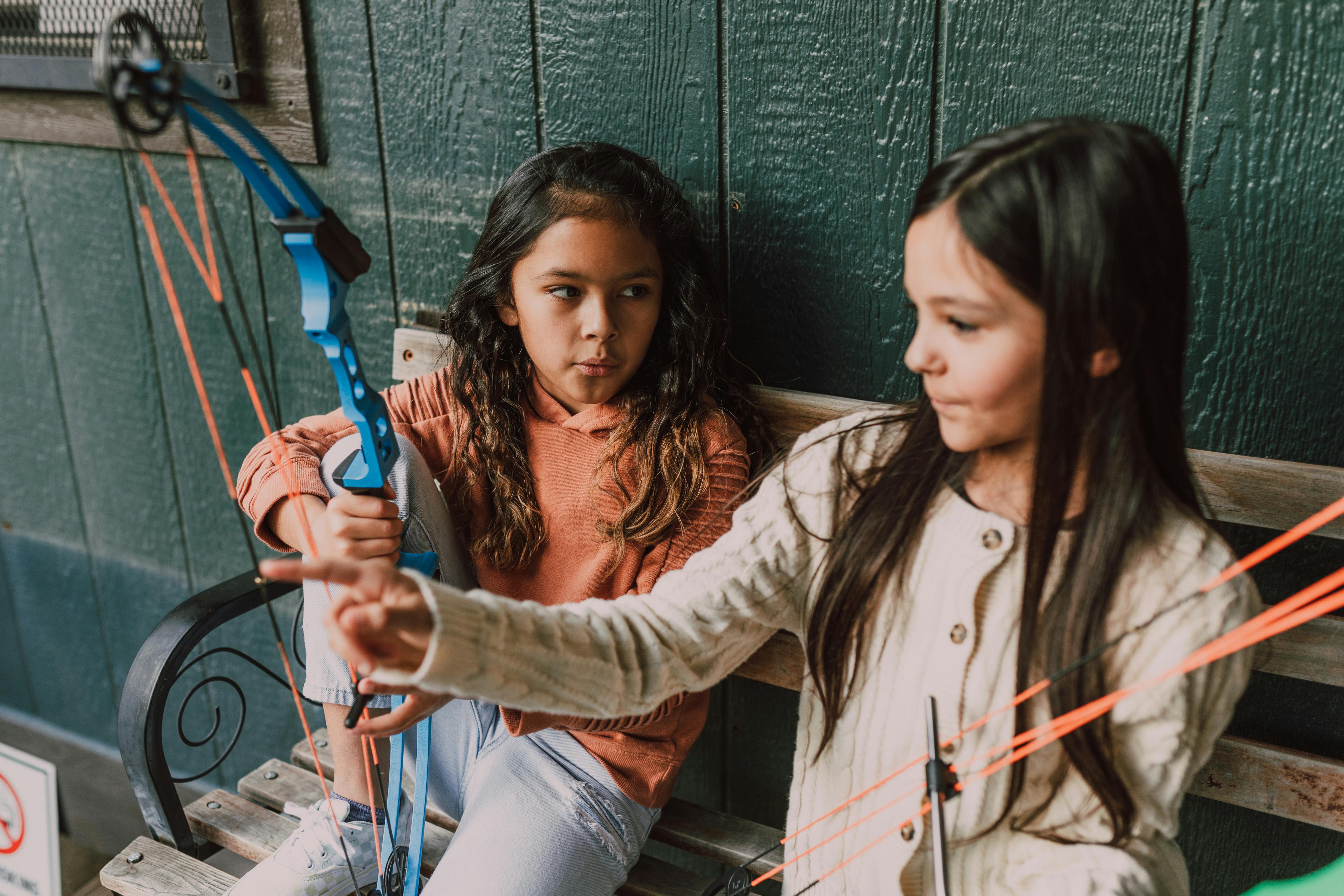 Two young girls engaged in archery practice with compound bows on a wooden bench indoors.