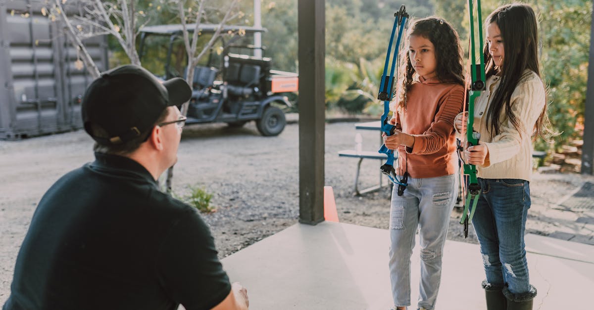 Two young girls learning archery with an instructor outdoors, focused and attentive.