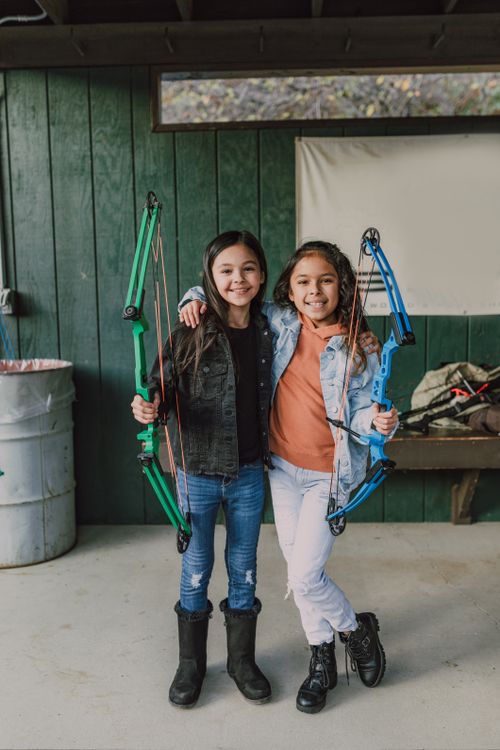 Free Two smiling girls indoors, holding colorful compound bows, enjoying archery practice. Stock Photo