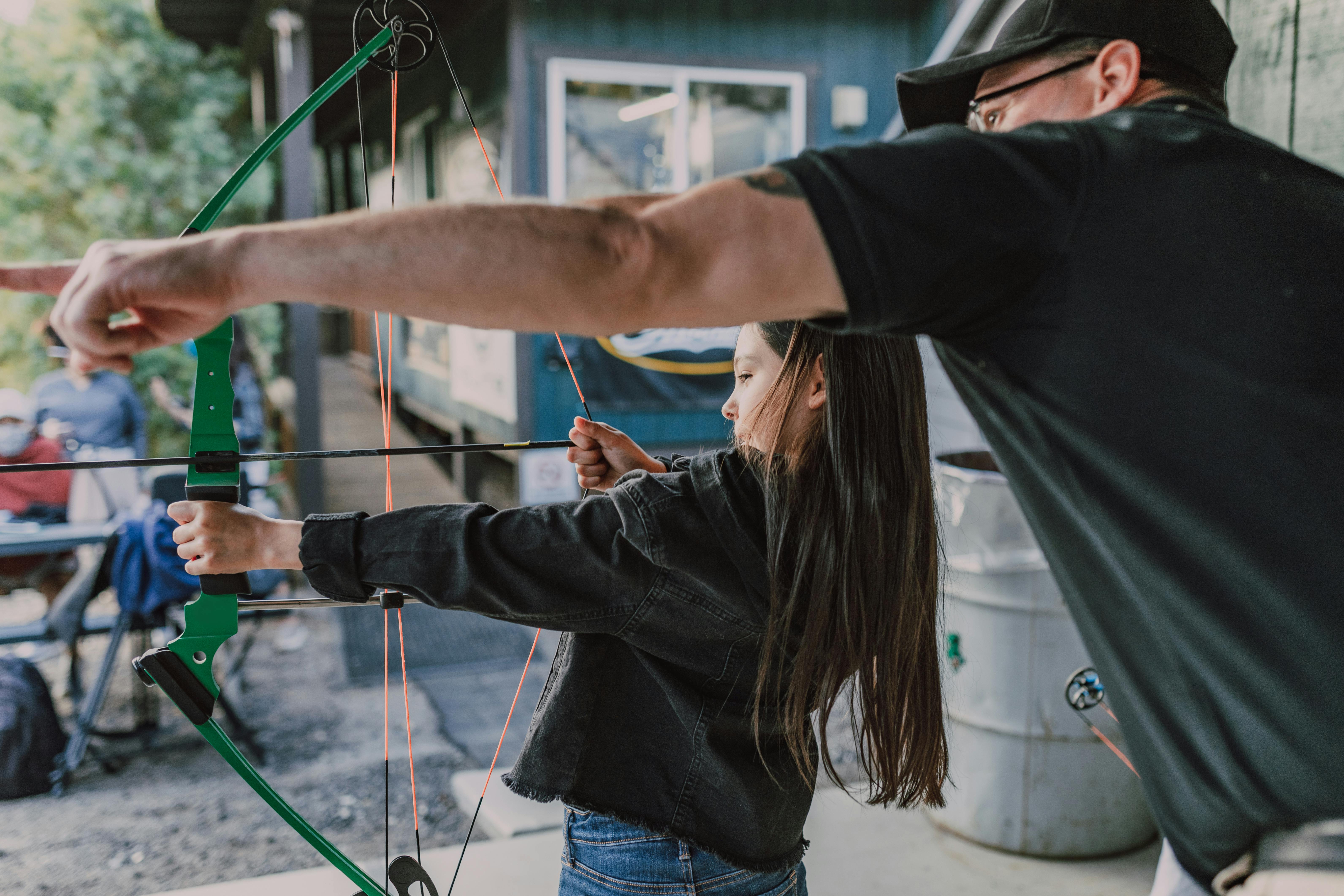 A young girl is learning archery with guidance from an adult man outdoors, focusing on target practice.
