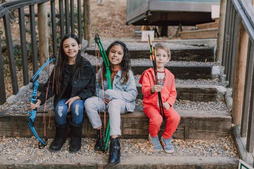 Free Three children with bows and arrows practicing archery outdoors, smiling and sitting on wooden steps. Stock Photo