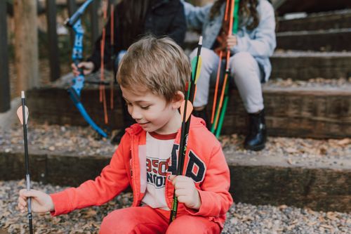 Free A young boy in a red outfit holds arrows happily while sitting outdoors. Engaging archery activity. Stock Photo