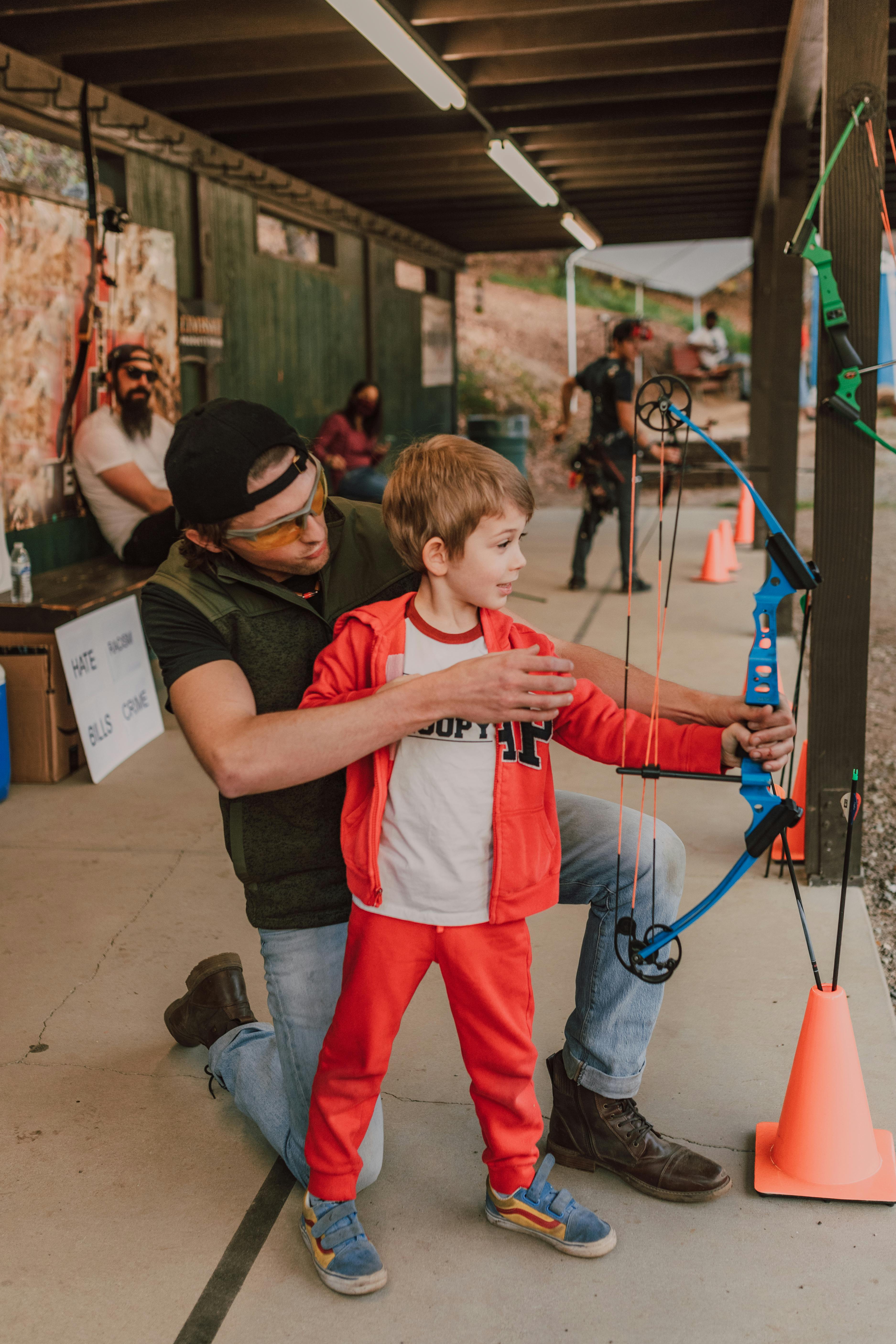 A father teaches his young son archery at an outdoor range, emphasizing skill and bonding.