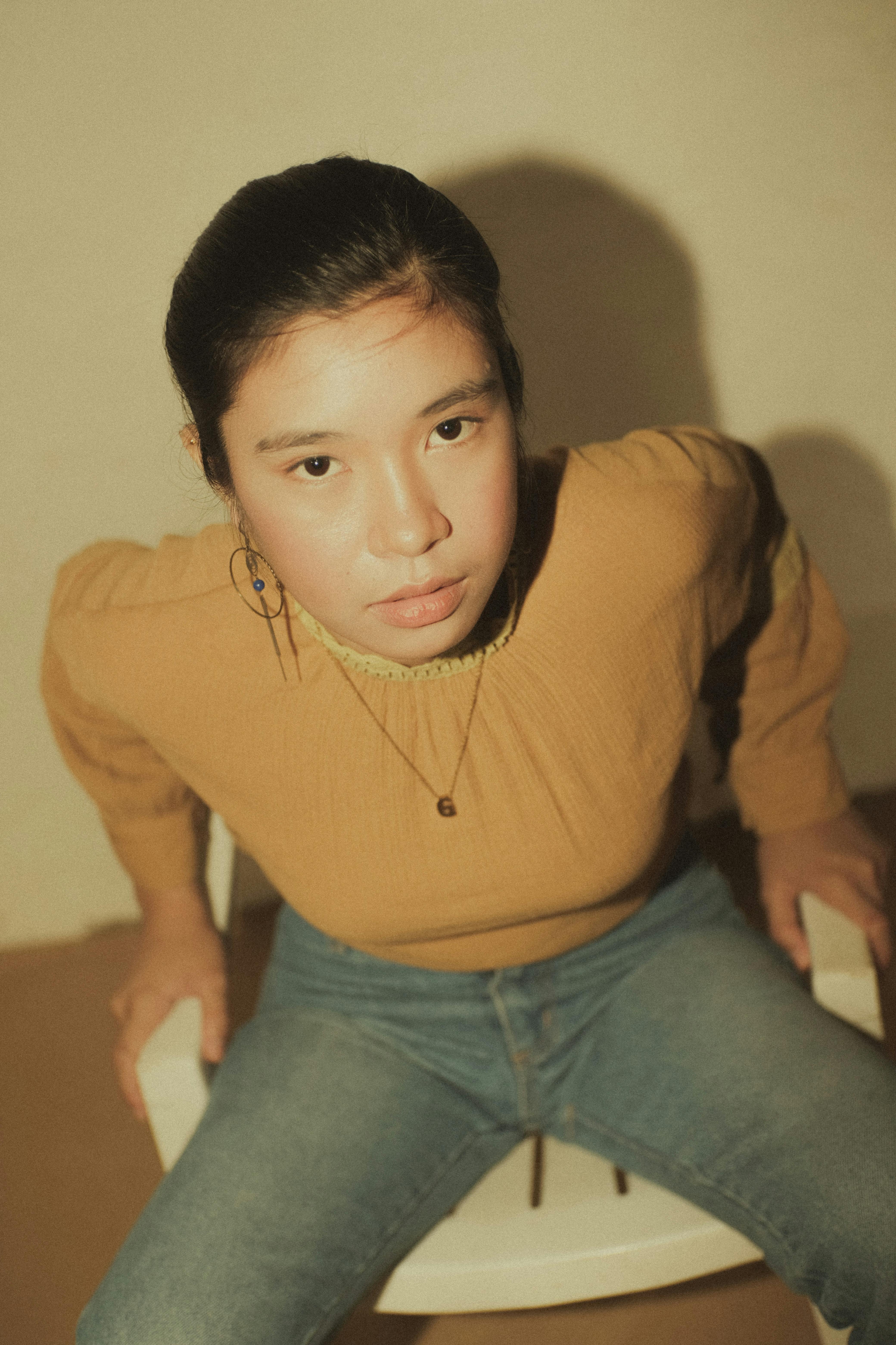 Serious young Asian lady in casual clothes and accessories sitting on plastic chair and looking at camera in light apartment
