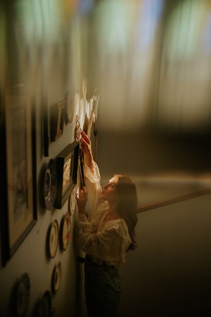 Young woman arranging picture frames on a well-decorated indoor wall with soft lighting.
