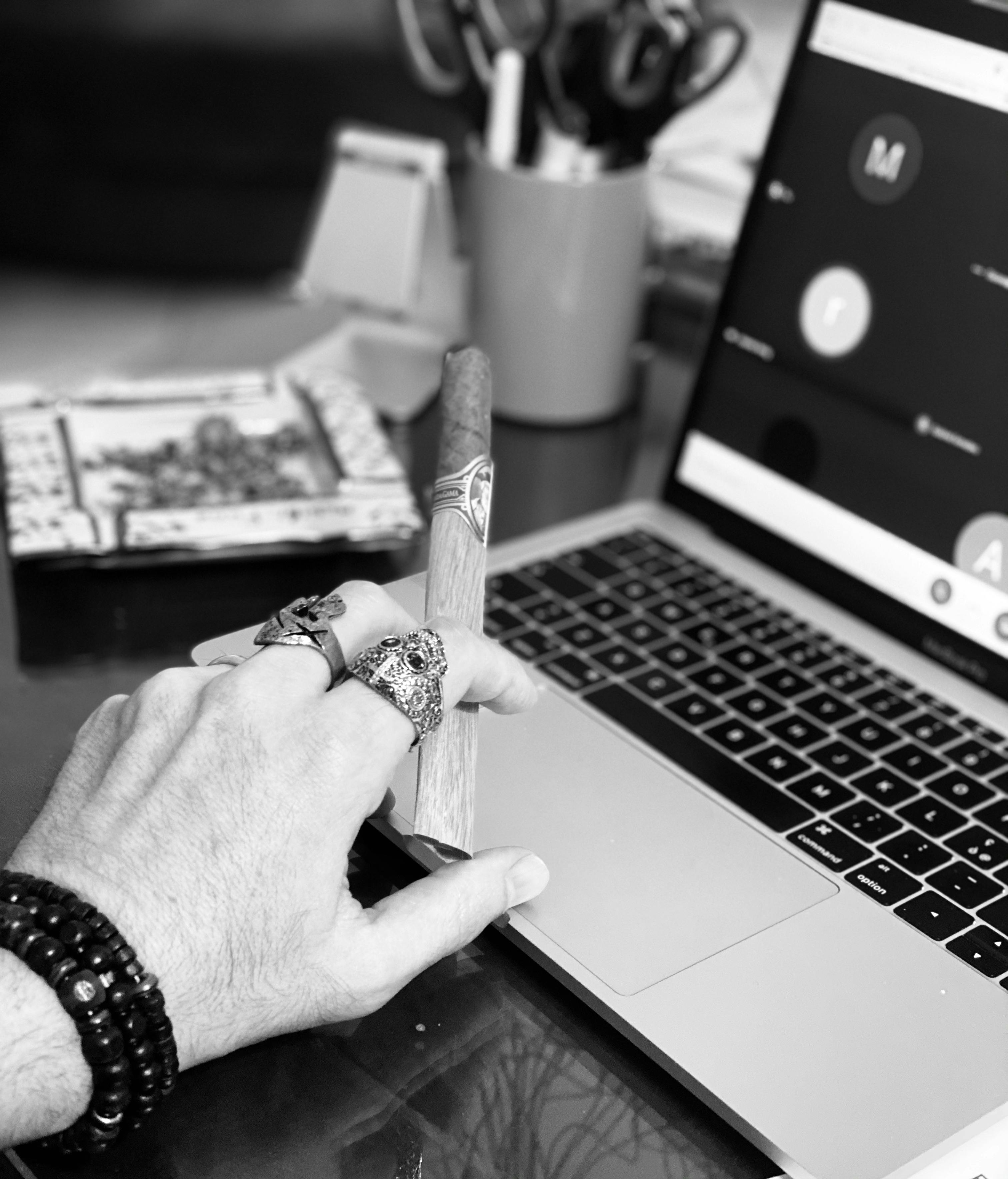 Black and white close-up of a man holding a cigar with rings, near a laptop on a desk.