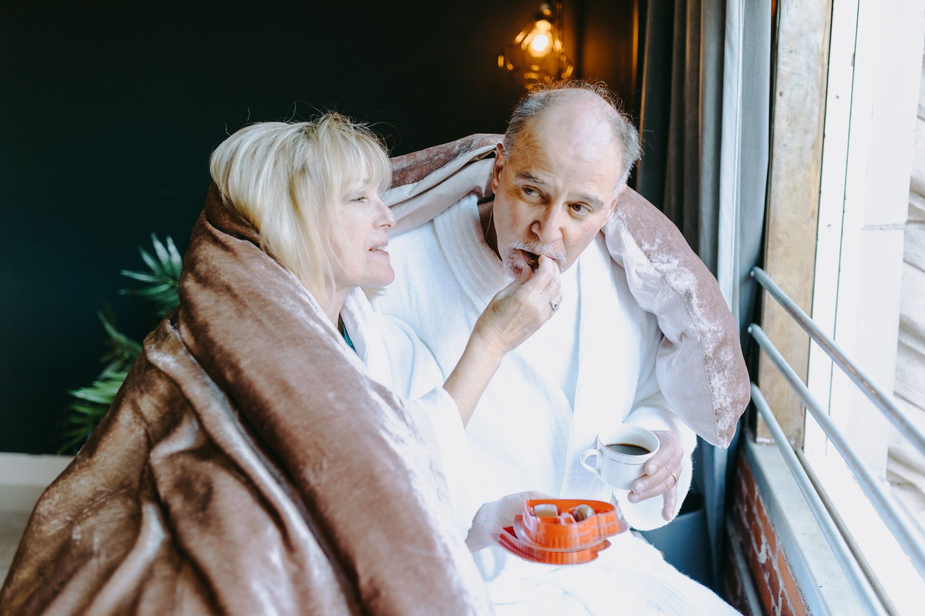 Warm moment shared by a senior couple in bathrobes, enjoying chocolates and a beverage by the window.