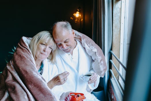 Senior couple shares a warm moment with coffee and chocolates, wrapped in a blanket by the window.