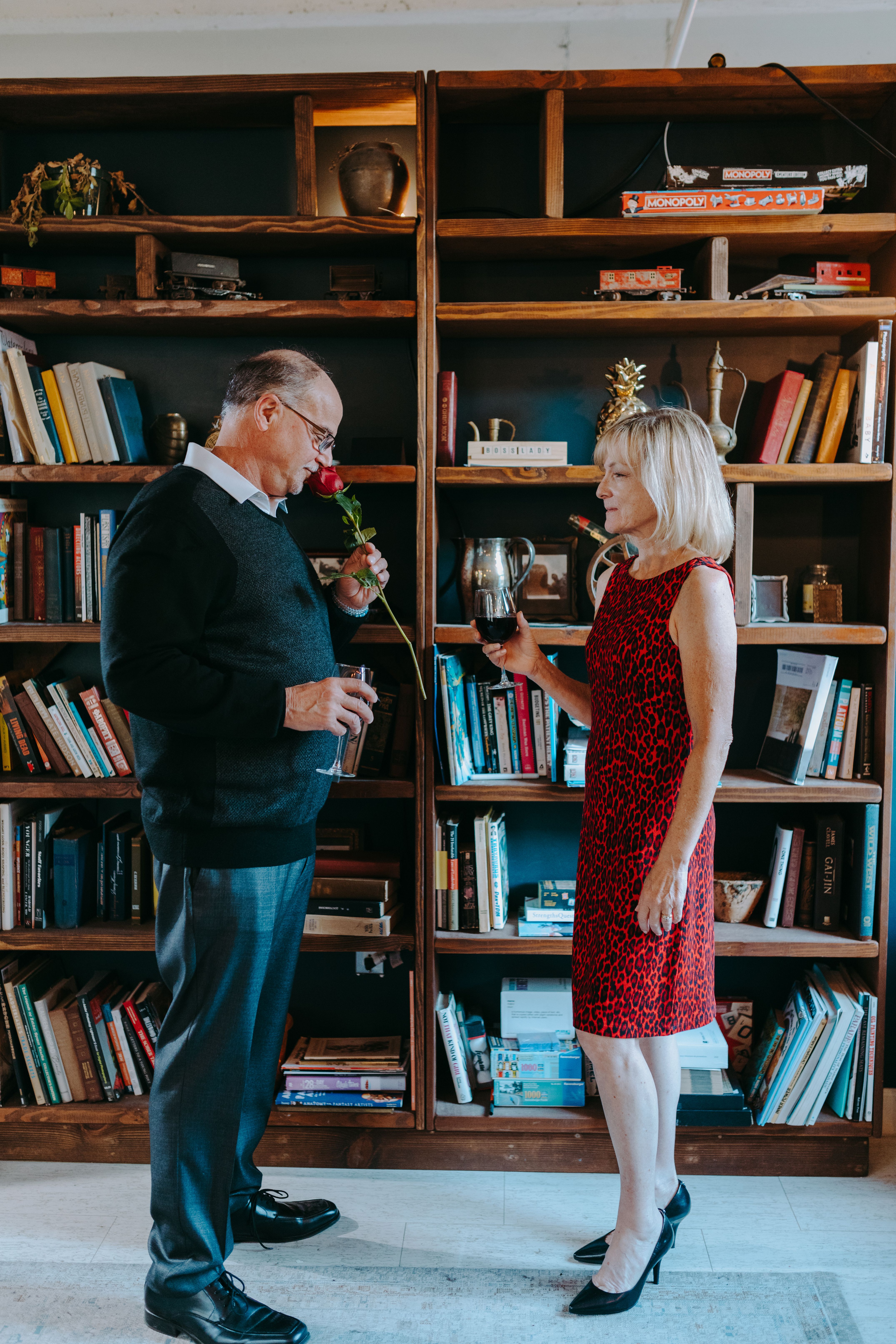 Elderly couple enjoying a romantic moment with wine and a rose in a cozy room with bookshelves.