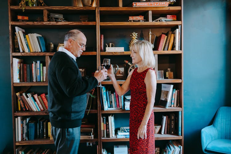 An Elderly Couple Toasting Drinks While Standing Near The Wooden Shelf