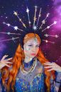 Self assured young female magician in dress with shining headband touching long red hair and looking away against purple starry sky
