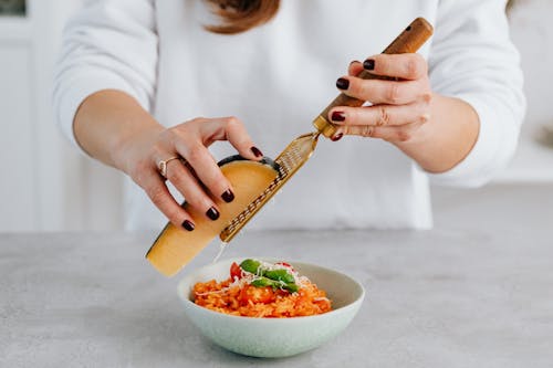 Free A woman with manicured nails grates parmesan over a tomato risotto, emphasizing culinary creativity. Stock Photo