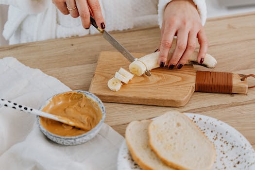 Female hand slicing banana on wooden board with peanut butter and bread for breakfast.