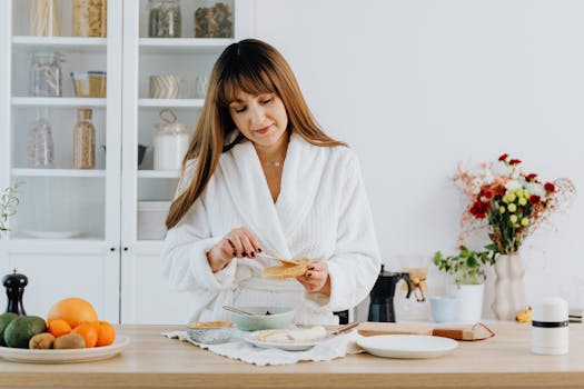 A woman in a bathrobe preparing breakfast in a home kitchen.