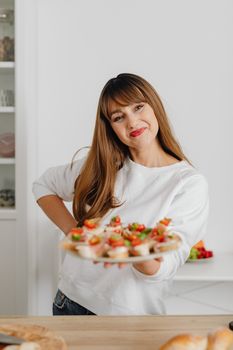 Smiling woman in a white top presenting a plate of colorful canapés indoors.