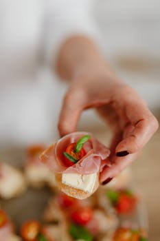 A hand holding a delicious gourmet cheese appetizer with tomato and ham.
