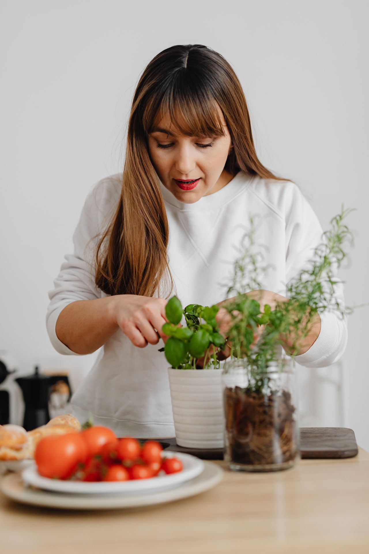Woman tending a potted basil plant on a kitchen counter next to rosemary growing in a glass jar