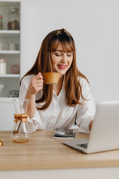 Free Woman in a white shirt enjoying coffee while working on a laptop at home. Stock Photo