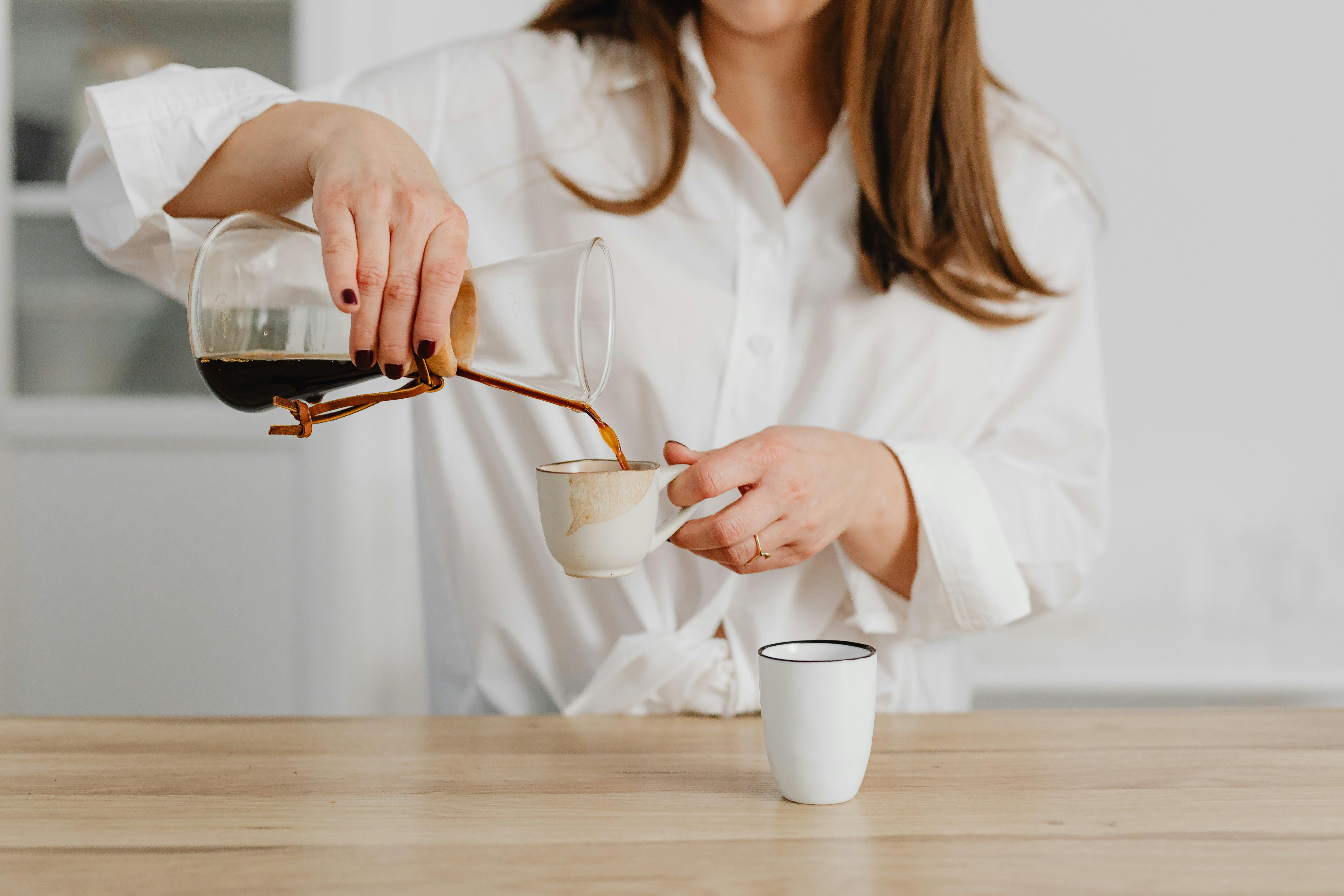 Woman in white shirt pouring coffee from a Chemex into a ceramic cup on a wooden table.