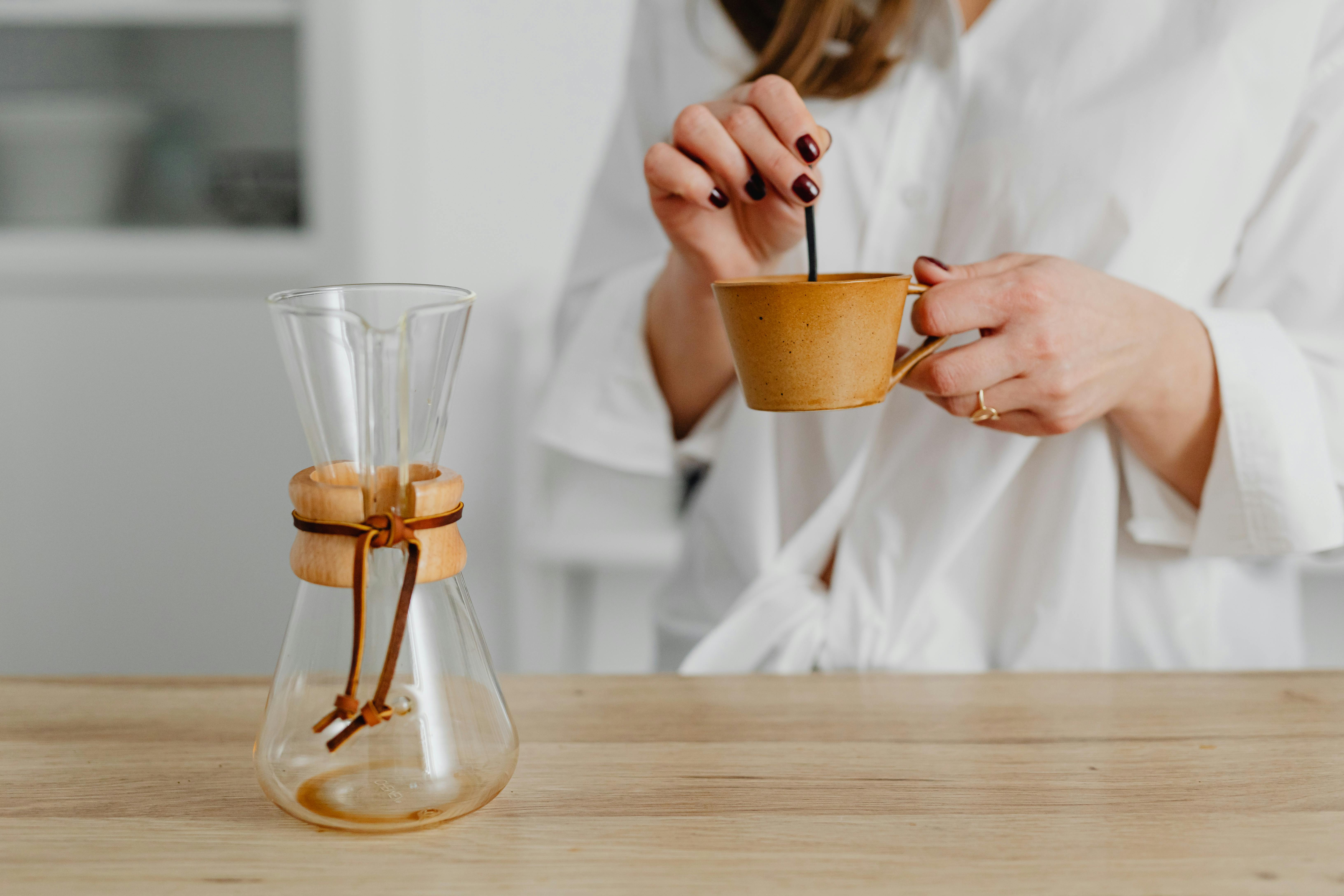 coffee brewing methods for home - Woman stirring coffee in ceramic cup beside blank Chemex on wood table.