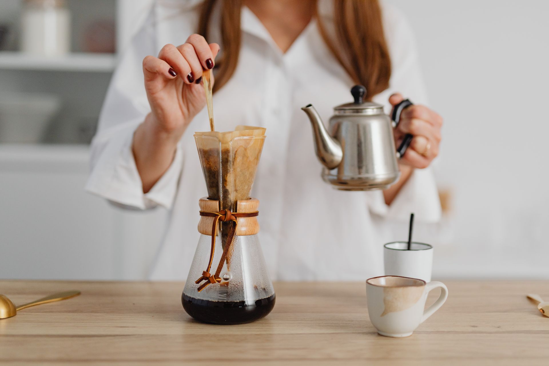 A woman brewing coffee using a pour-over method with a Chemex and kettle.