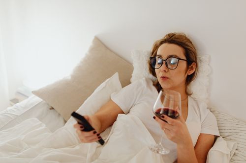 Free A woman wearing glasses relaxes in bed, holding a wine glass and remote control. Stock Photo