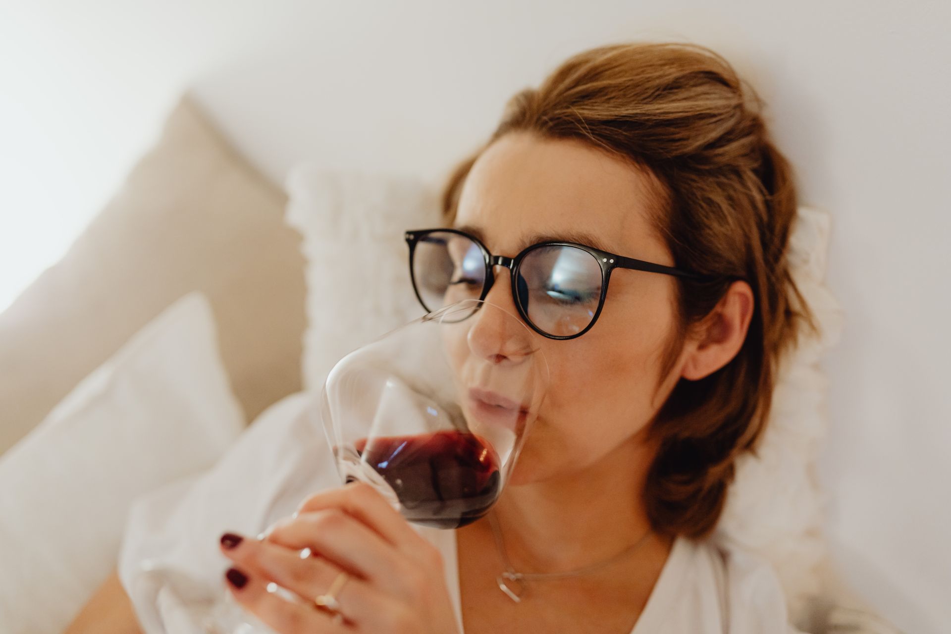 A woman in glasses relaxes indoors, sipping red wine peacefully.