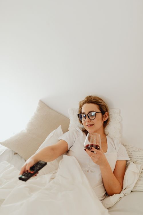 Free Cozy scene of a woman lying in bed, holding a wine glass and a remote control. Stock Photo