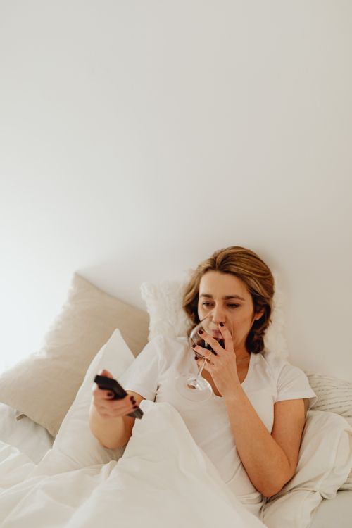 Free A woman lies in bed enjoying a glass of wine and watching TV with a remote. Stock Photo