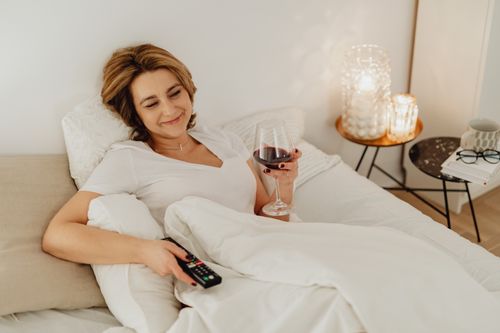 Free Woman enjoying a relaxing evening in bed, sipping red wine while watching TV in a cozy bedroom setting. Stock Photo