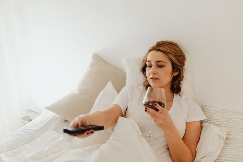 Free Woman in bed holding wine glass and remote, enjoying a cozy, relaxing moment. Stock Photo