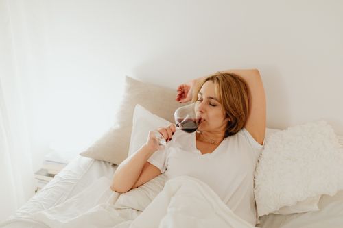 Free Woman in white shirt relaxing in bed, sipping red wine with pillows and soft lighting. Perfect for lifestyle themes. Stock Photo