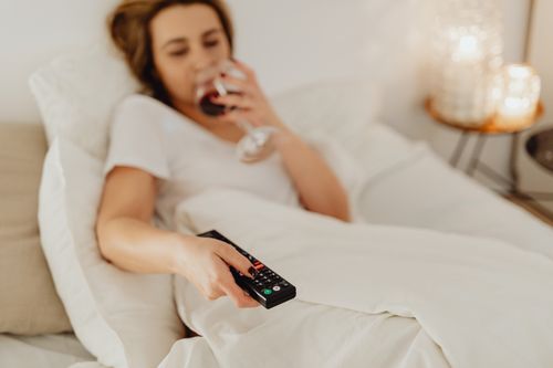Free A woman in bed holding a glass of wine and a remote control, enjoying a cozy evening. Stock Photo