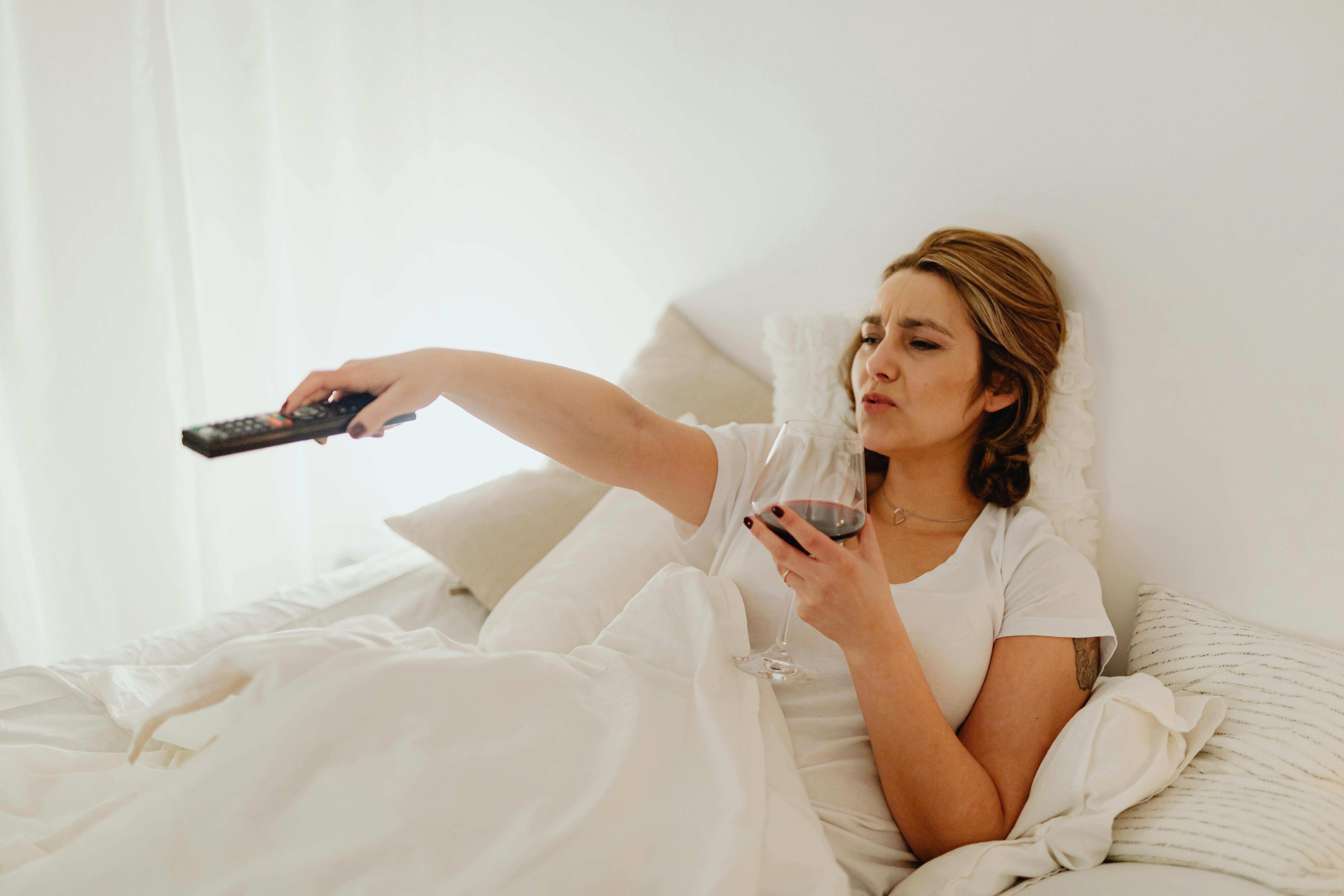 A woman in bed enjoying a glass of red wine while holding a TV remote, creating a relaxed atmosphere.