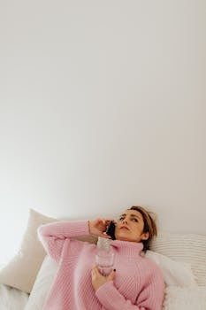 Woman in pink sweater lying in bed, talking on mobile and holding a glass of water in bright bedroom setting.