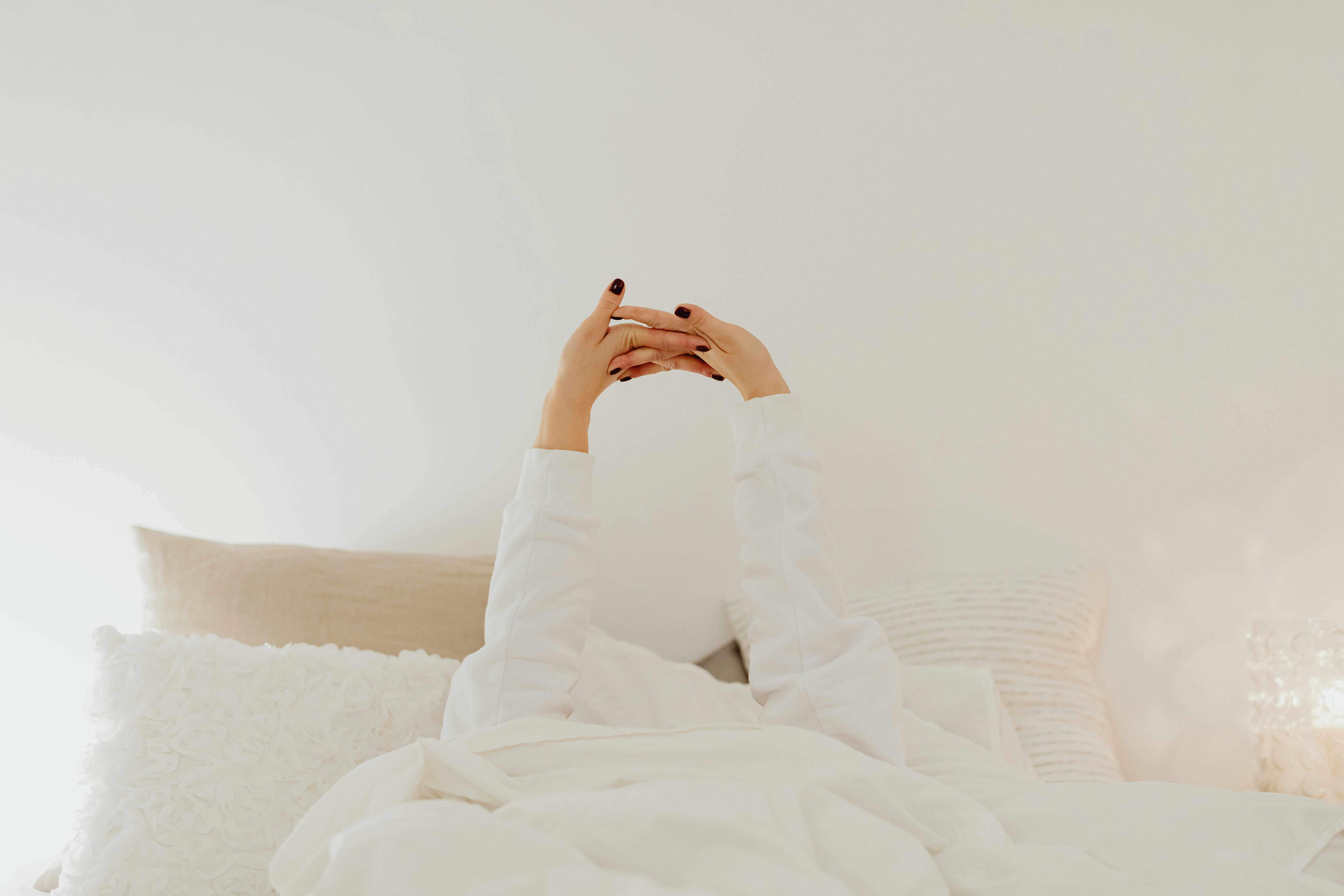 A person stretches in bed, surrounded by soft white linens in a calming bedroom.