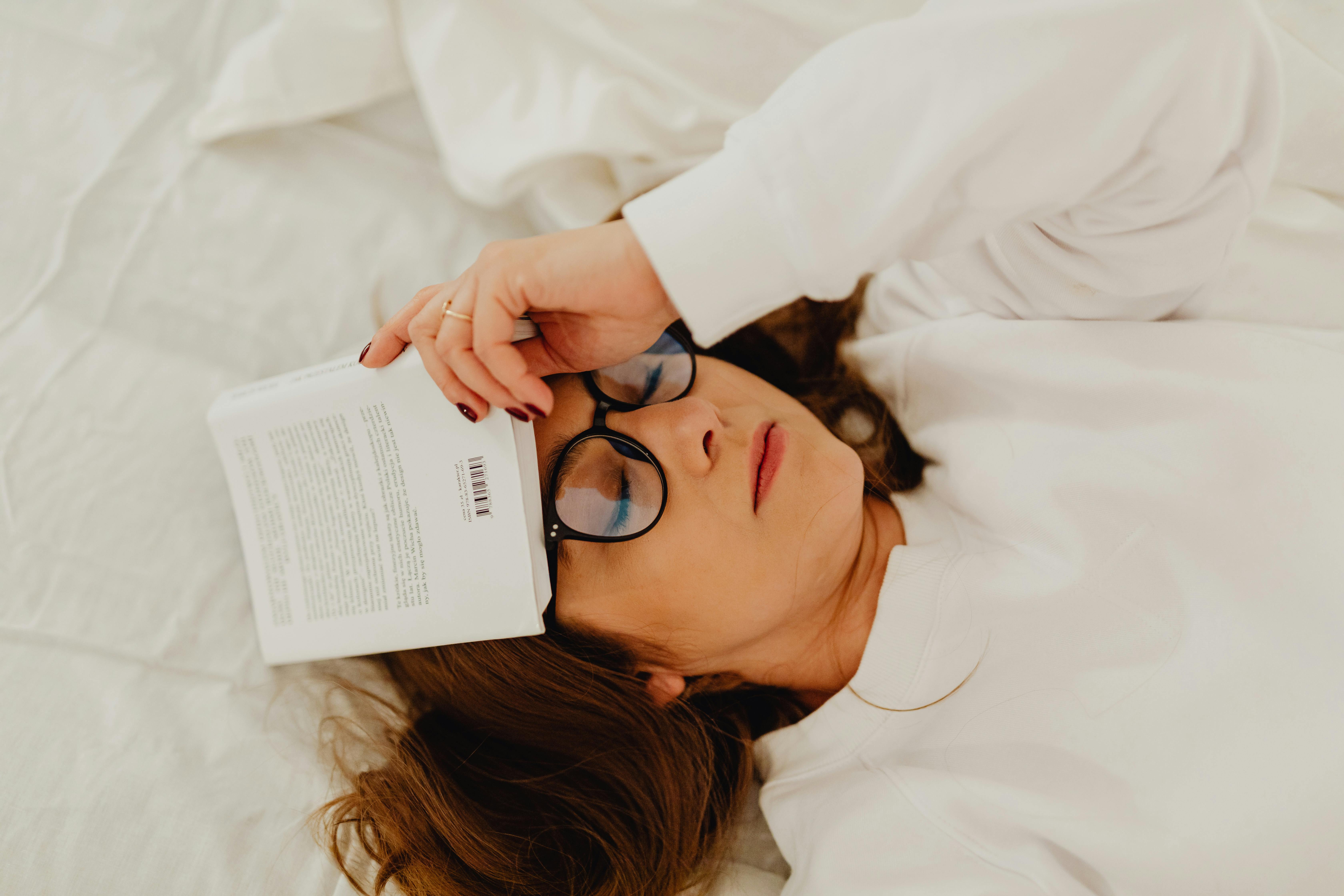 Woman lying in bed with a book over her face, wearing glasses and a cozy sweater.