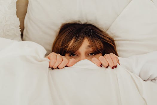 A woman with manicured nails peeks over a white blanket in a cozy setting.