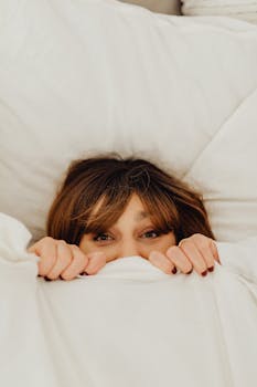 A woman playfully peeks out from under a white blanket in a cozy bedroom setting.