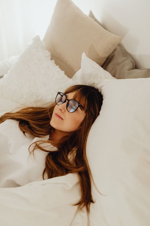 Free A peaceful woman with eyeglasses resting on a bed with fluffy pillows in a serene bedroom setting. Stock Photo