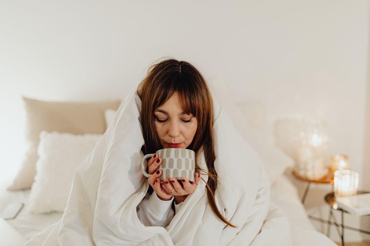 Woman wrapped in a white throw drinking tea by candlelight